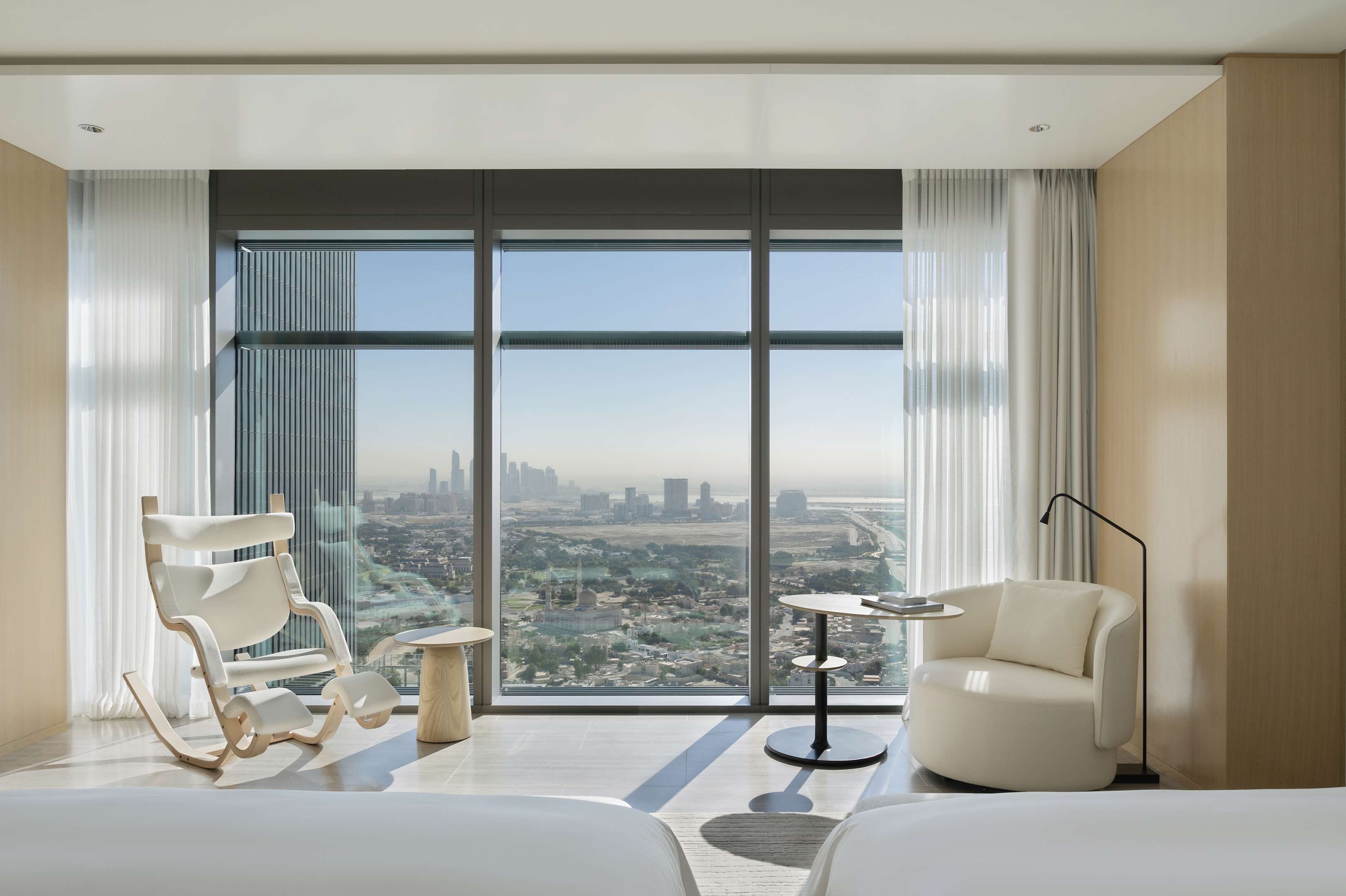 A modern hotel room with a large window offering a city view, a white sofa, a small round table, and a bed.