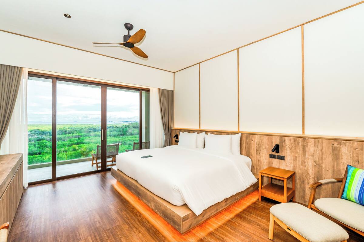 Una habitación de hotel moderna con una cama grande, piso de madera, ventilador de techo, una mesita de noche de madera, una silla y vistas a un paisaje verde a través de un ventanal amplio.
