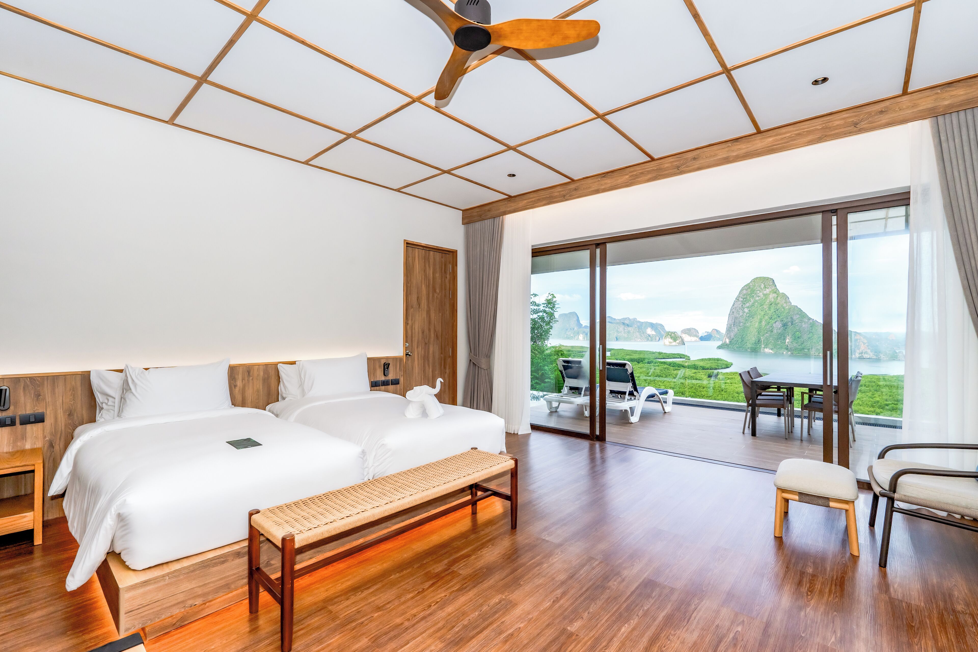Habitación de hotel moderna con una cama grande, un banco, un comedor con asientos al aire libre y una vista panorámica de una montaña y un lago.