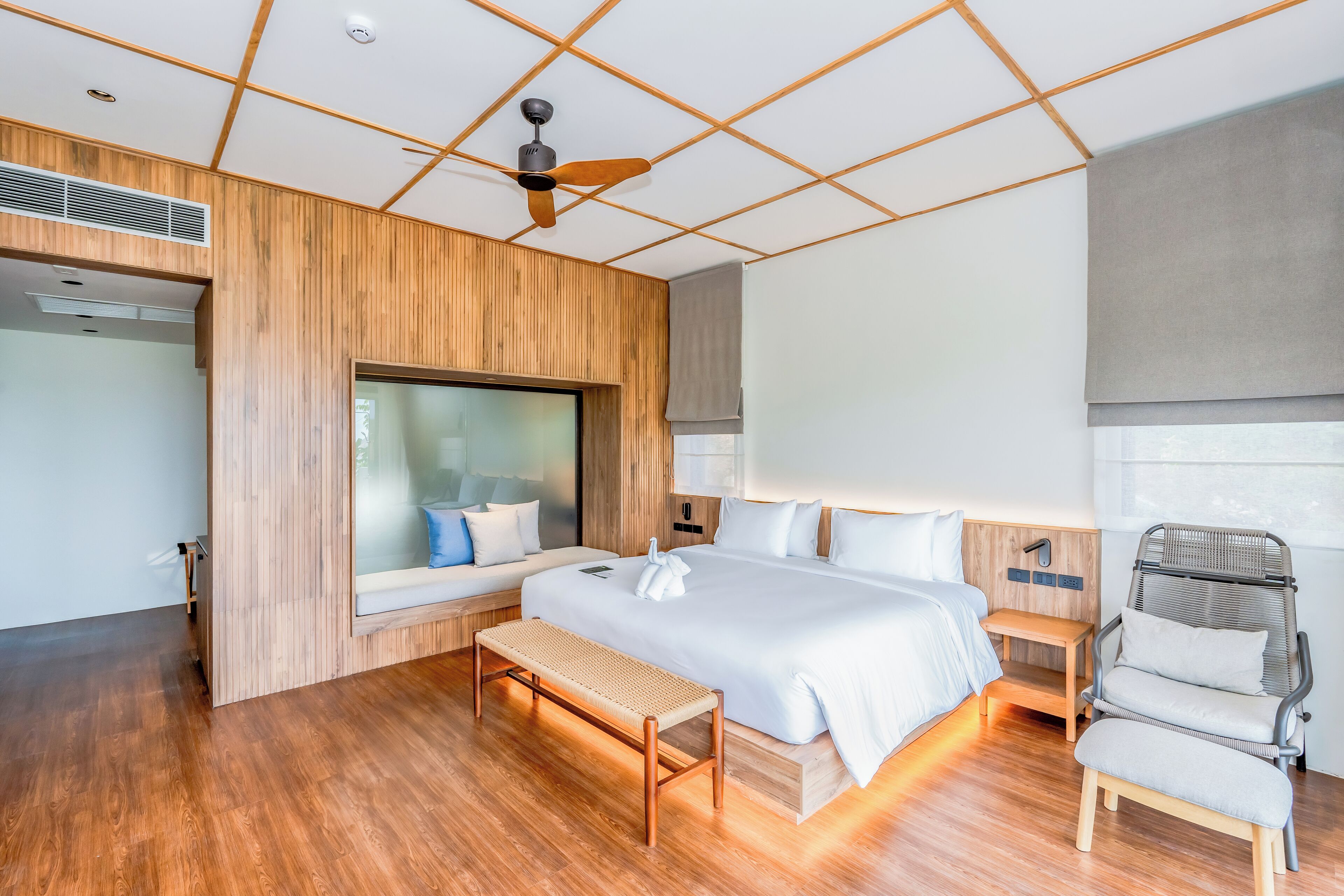 Ein modernes Hotelzimmer mit einem großen Bett, einer hölzernen Bank, einem Deckenventilator und einem großen Fenster.