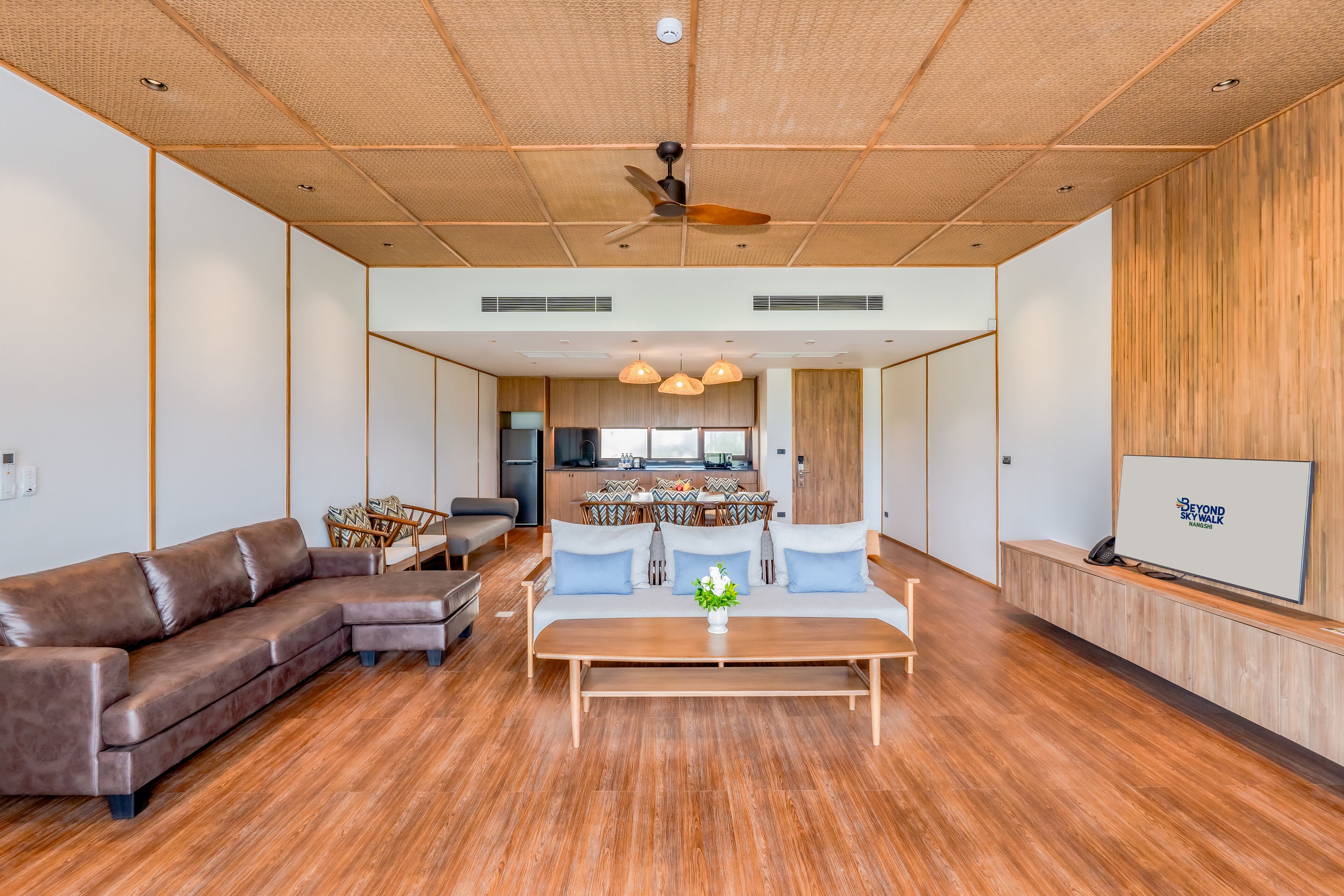 Un salón espacioso con suelo de madera, un sofá de cuero marrón, una mesa de centro de madera y una zona de comedor con una cocina al fondo.