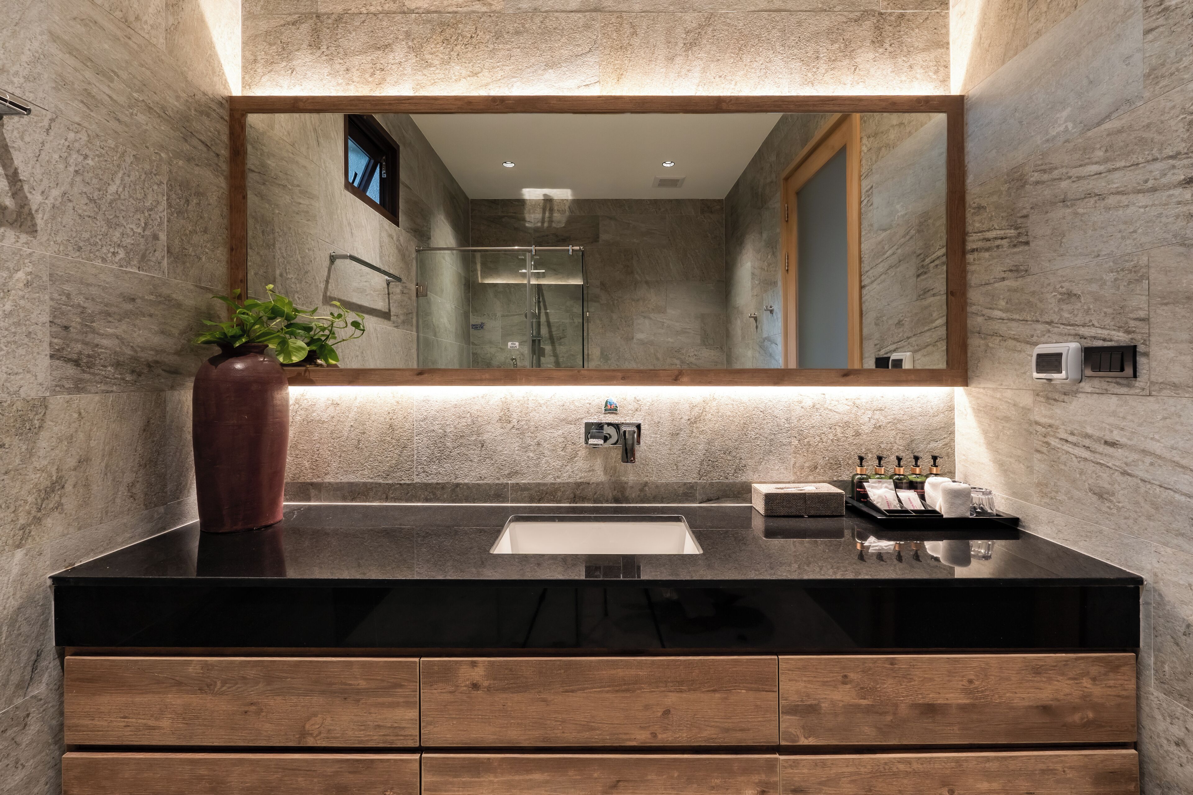 Un baño moderno con encimera negra, armarios de madera, un espejo grande y zona de ducha.