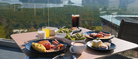 Una mesa con alimentos para el desayuno y un periódico, con vistas a un paisaje pintoresco que incluye montañas y un puente.