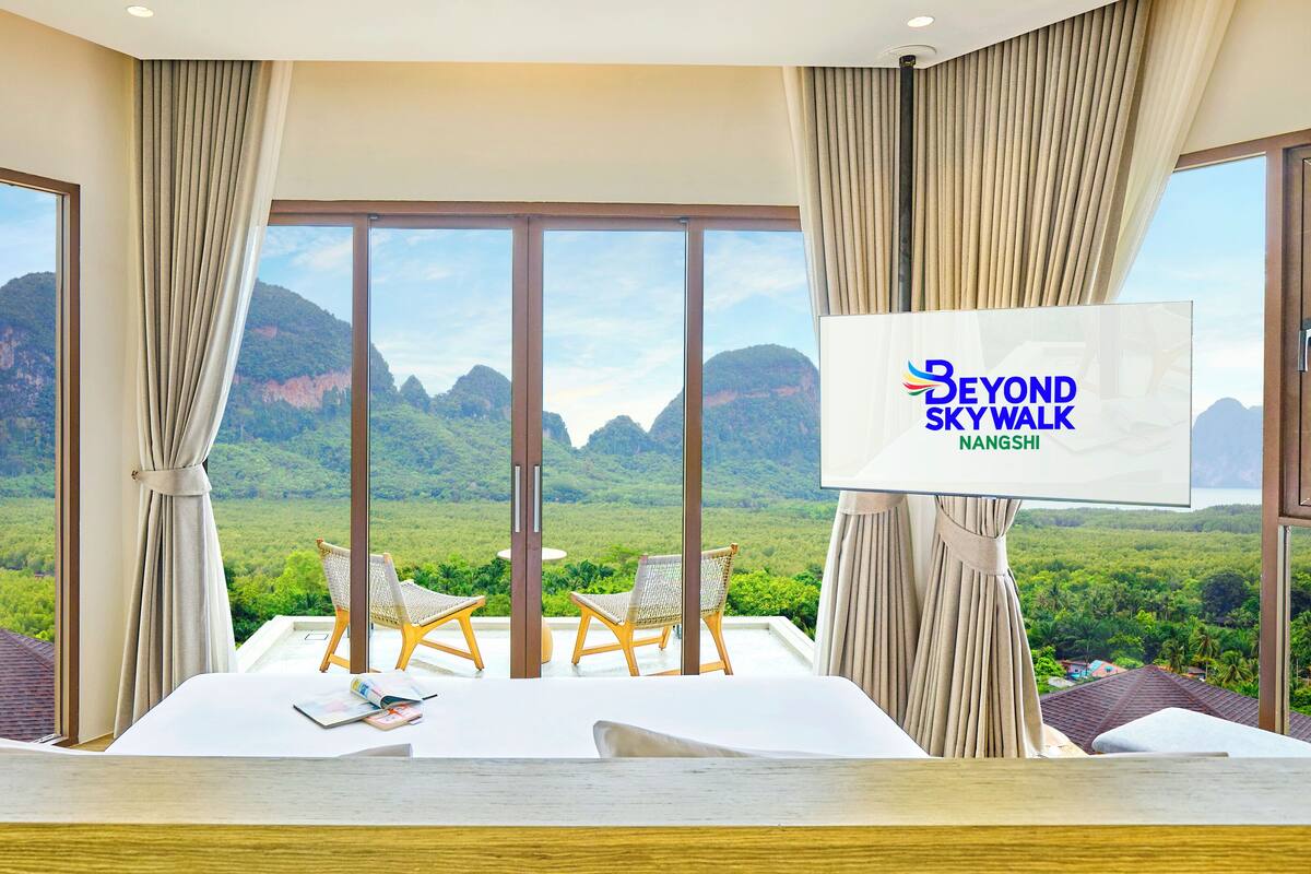 Habitación de hotel con un ventanal grande que ofrece vistas a un paisaje montañoso. Hay una cama con sábanas blancas, dos sillones de mimbre y un televisor de pantalla plana montado en la pared.