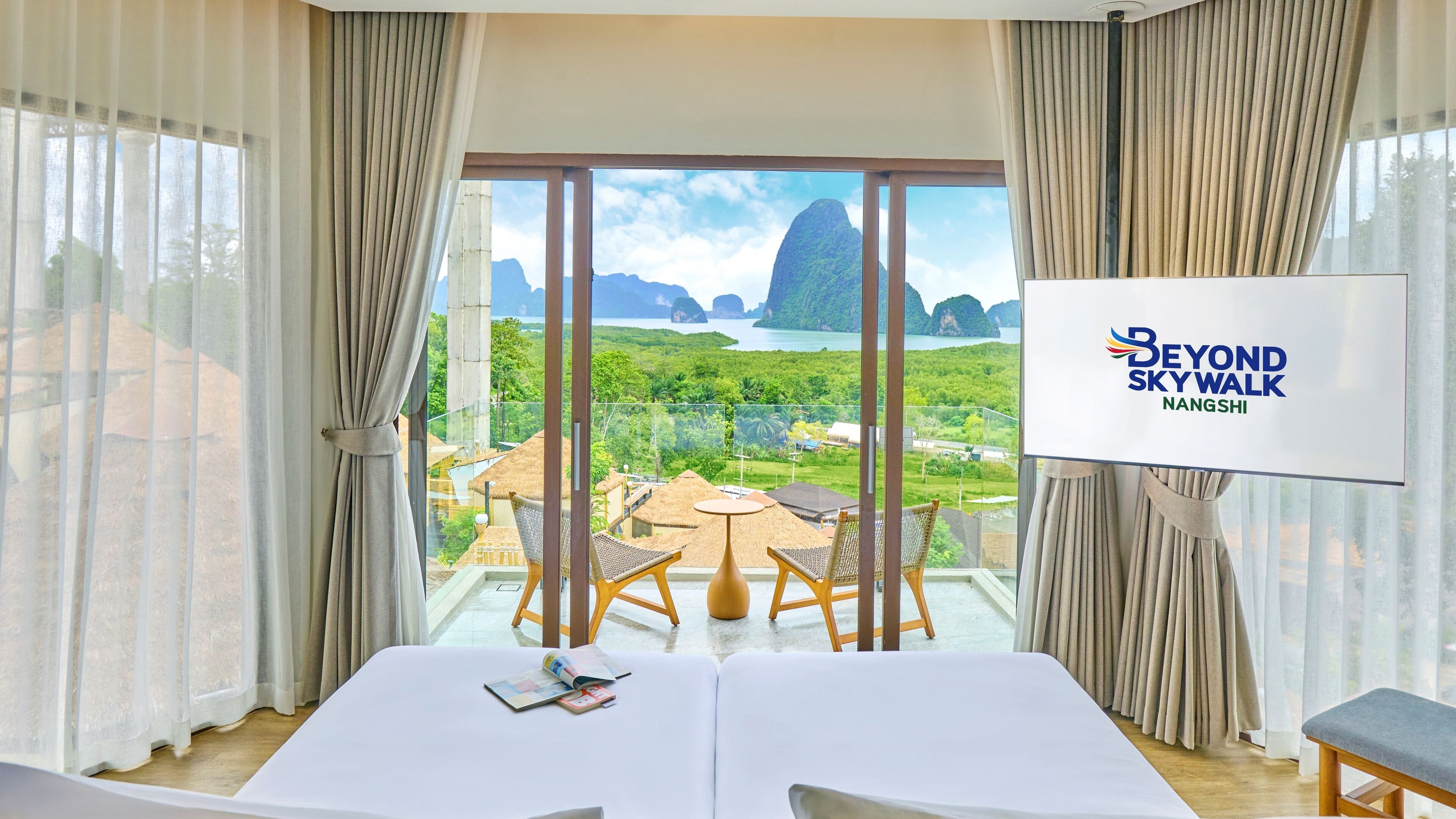 A hotel room with a large window offering a view of a lush landscape and mountains. There is a bed, a small table, and two chairs. A sign on the wall reads "Beyond Sky Walk Nangshi".