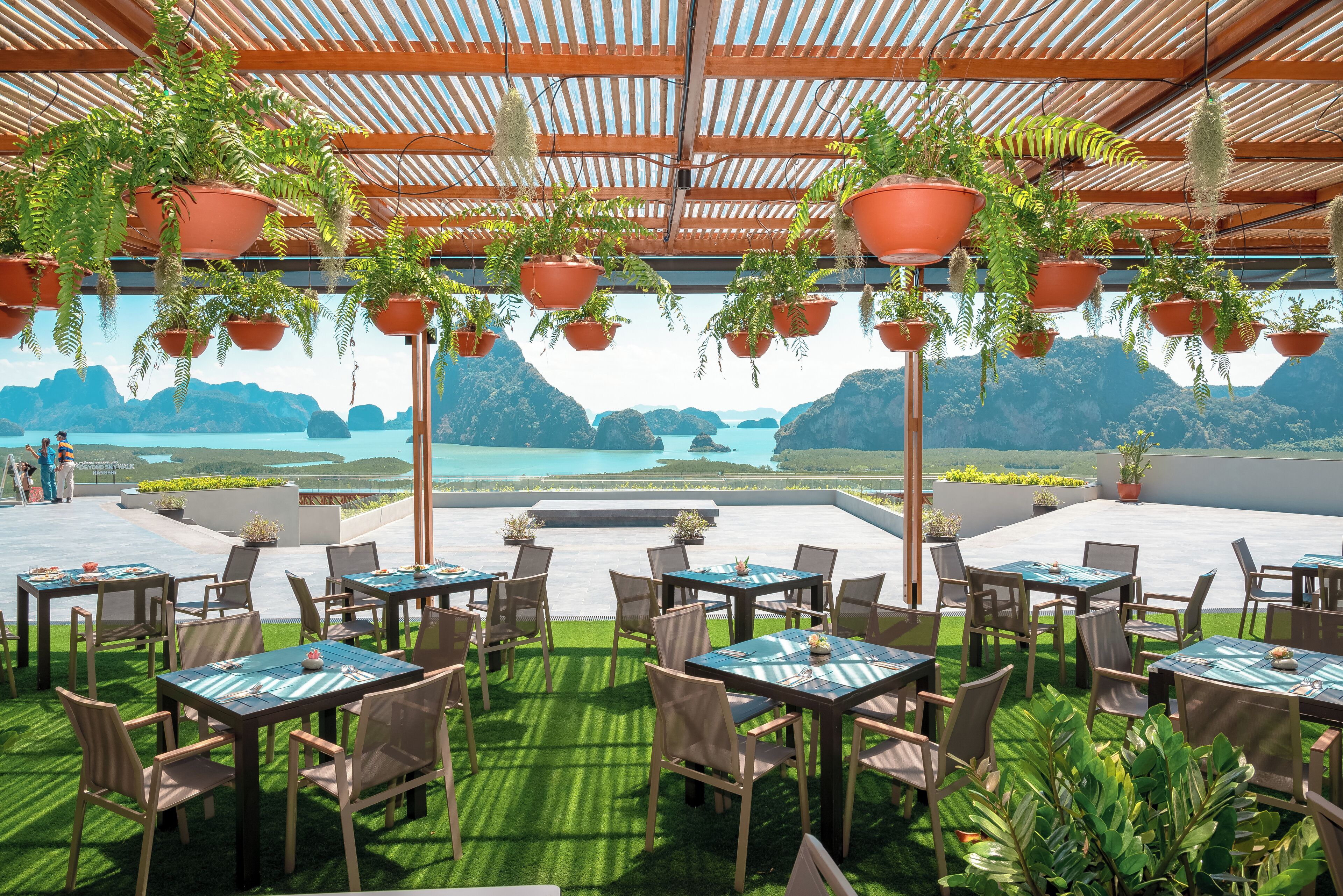 Un espace de restauration en plein air, avec des tables et des chaises, entouré de jardinières suspendues et offrant une vue sur une étendue d’eau.