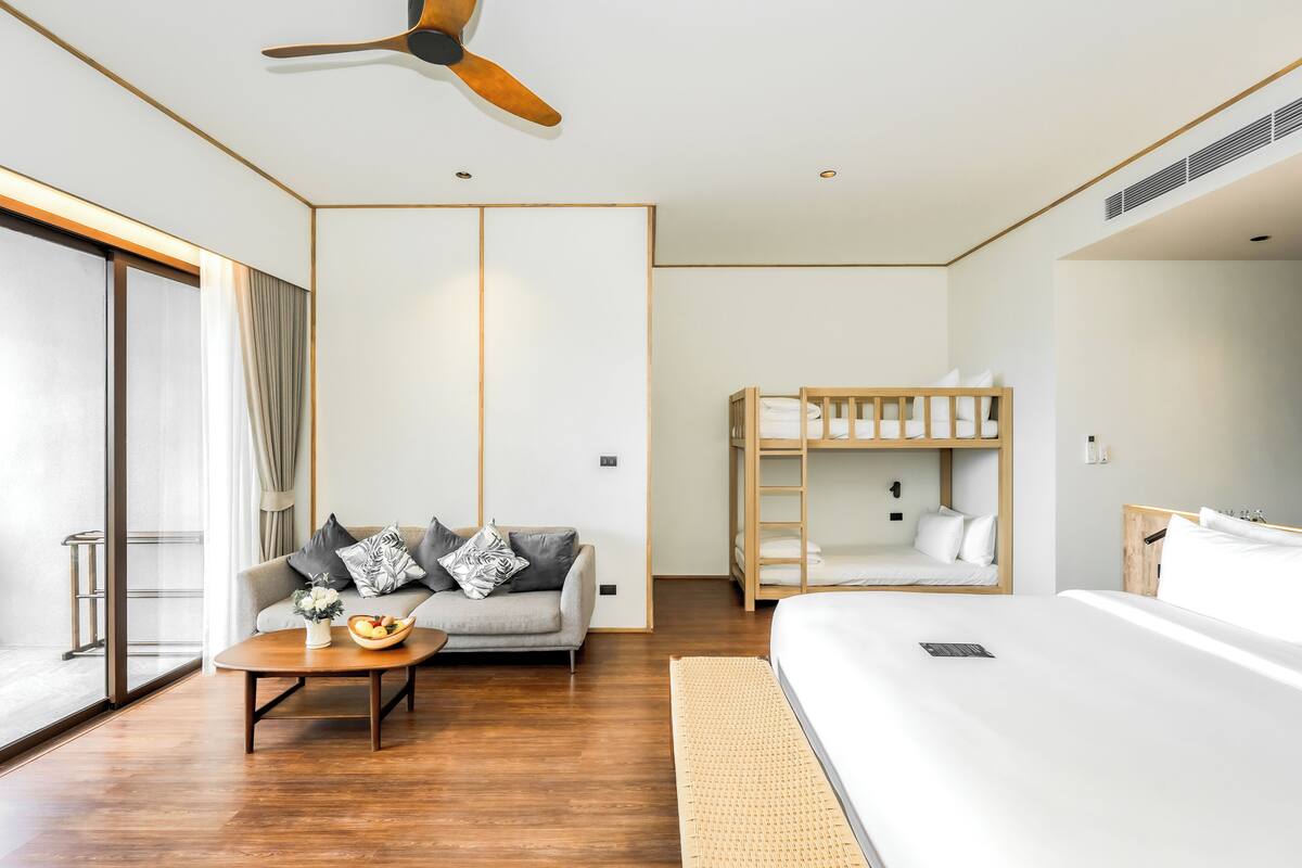 Una habitación de hotel moderna con una cama grande, un sofá, una mesita con un frutero, un ventilador de techo y una cama litera.