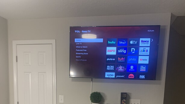 A flat-screen TV mounted on a wall displaying a Roku TV interface with various streaming options. - Welcome Home (Charlotte)