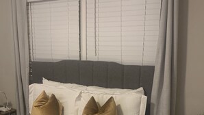 A neatly made bed with white bedding and two gold pillows, a grey headboard, and a window with blinds. - Welcome Home (Charlotte)