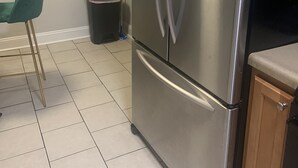 A stainless steel refrigerator in a kitchen with tiled flooring, a green chair, and a trash bin. - Welcome Home (Charlotte)