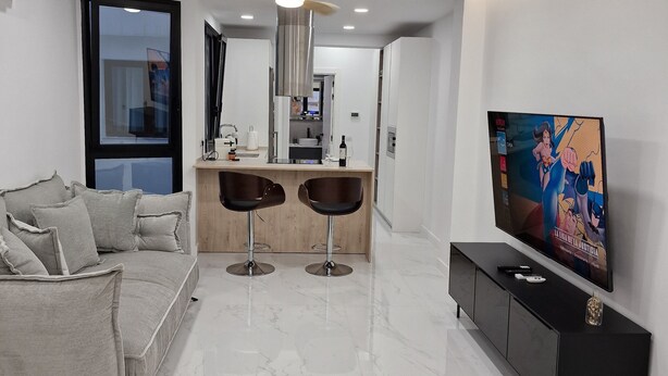 Uma sala de estar moderna com um sofá, dois bancos altos, uma televisão e uma cozinha ao fundo.