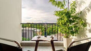 A balcony with a table, two chairs, a potted plant, and a view of greenery and a building in the distance.