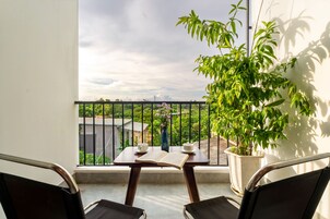 A balcony with a table, two chairs, a potted plant, and a view of greenery and a building in the distance.