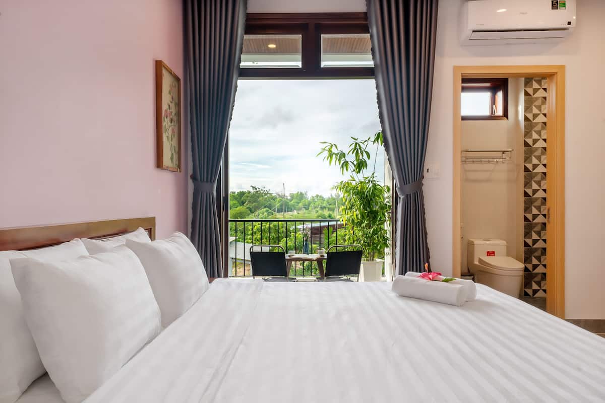 A hotel room with a large bed, a balcony with a table and chairs, and a view of greenery.