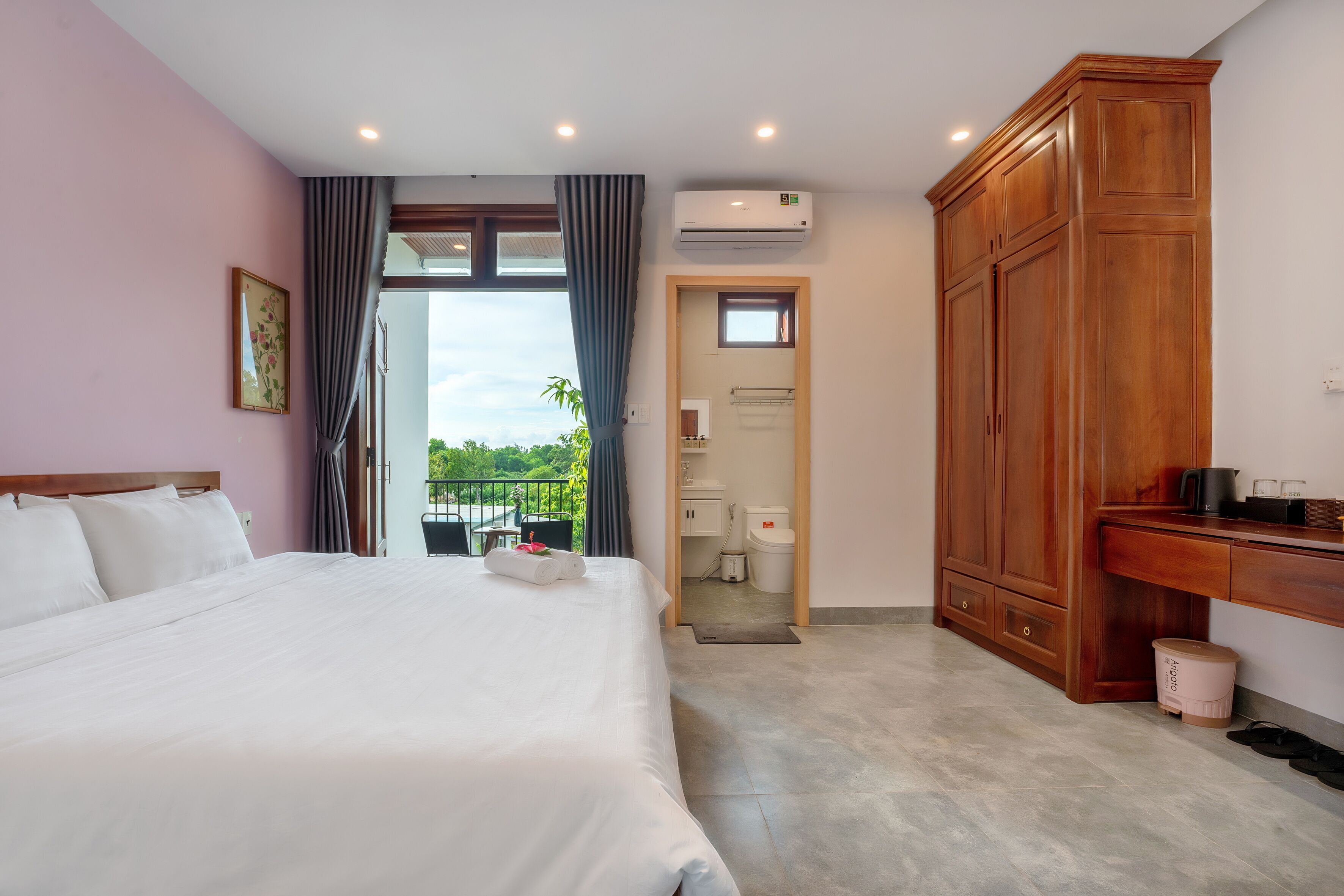 A hotel room with a large bed, a wooden wardrobe, an air conditioning unit, and a view of the outdoors.