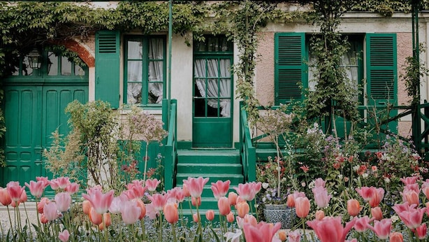Un bâtiment avec des volets verts et un jardin où poussent des fleurs roses.