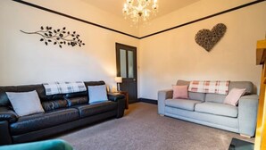 Un salon avec un canapé en cuir foncé, un canapé gris clair et un fauteuil turquoise. Un lustre est suspendu au plafond et une décoration en forme de cœur orne le mur.