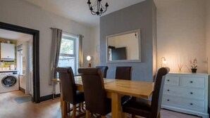 Un coin repas avec une table et des chaises en bois, un lustre, un miroir au mur et une commode blanche.