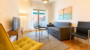 Ein Wohnzimmer mit einer Couch, einem gelben Sessel, einem Fernseher, einer Lampe und Blick ins Freie.