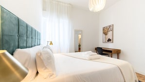 Ein ordentlich bezogenes Bett mit Kopfteil, ein Schreibtisch mit Stuhl und ein an der Wand hängendes Bild.