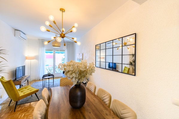 A dining area with a wooden table, chairs, a vase with flowers, a chandelier, and a wall-mounted mirror.