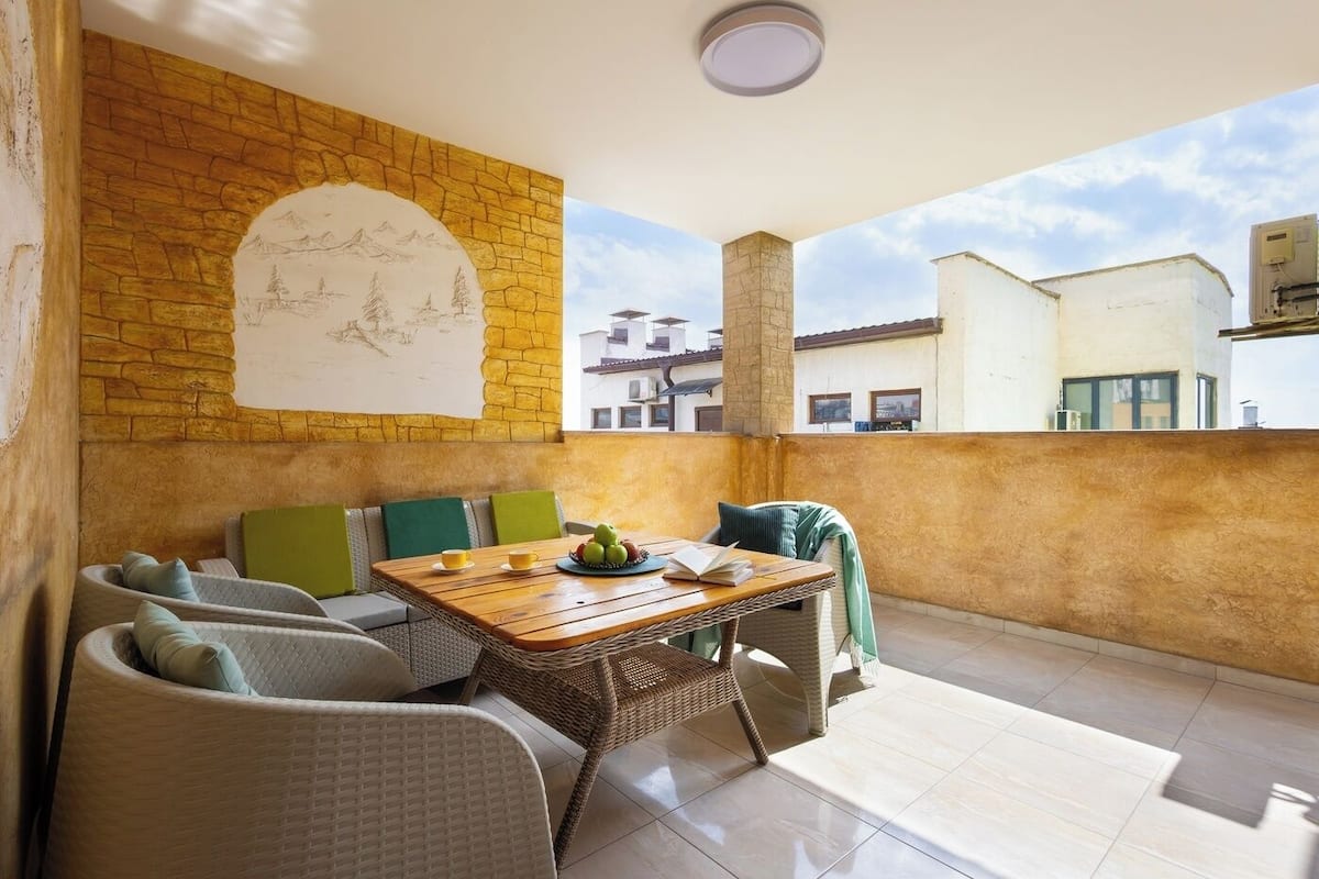 A balcony with wicker furniture, a table with a fruit bowl and book, and a mural on the wall.