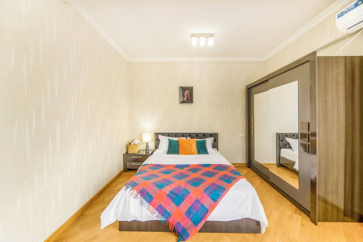 A hotel room with a bed, a nightstand, a mirror, and a wall-mounted air conditioning unit.