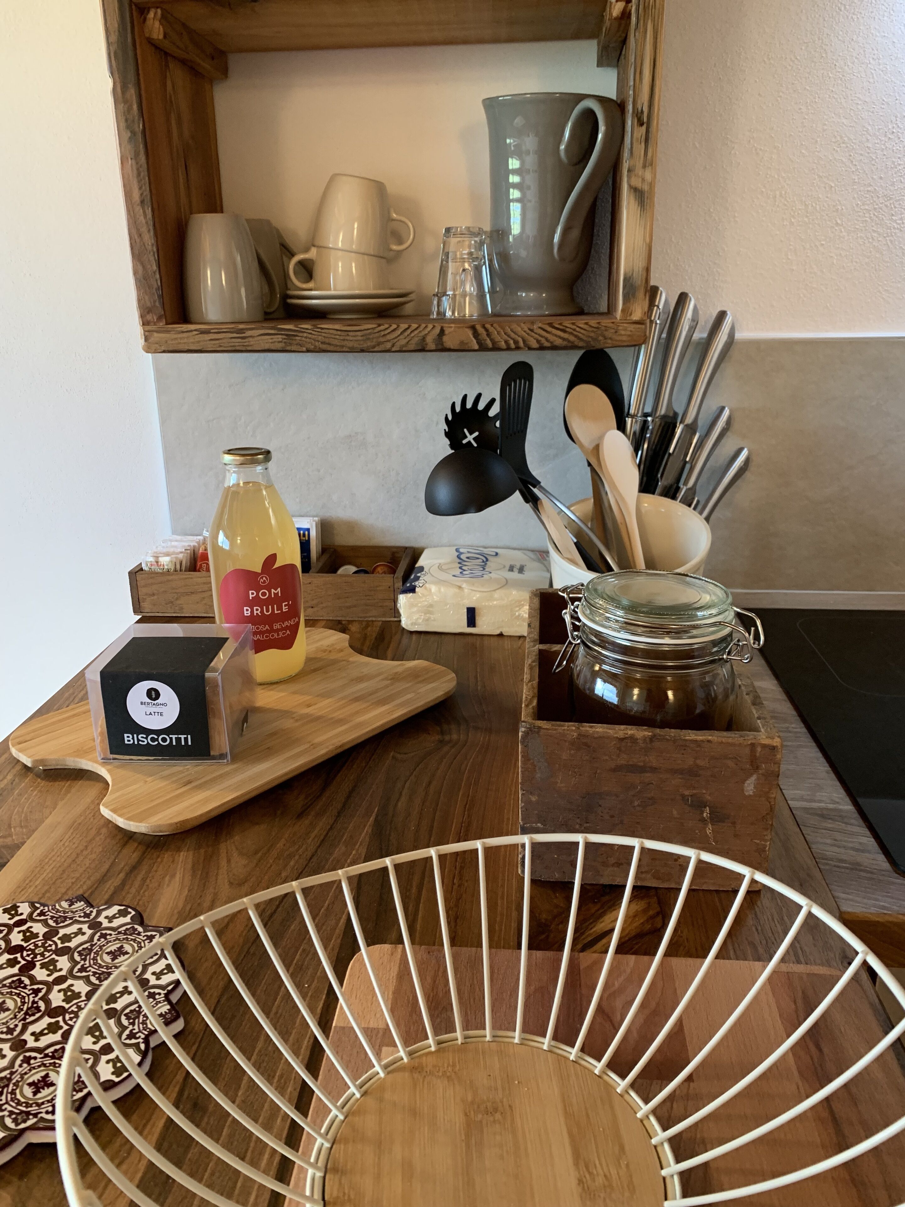 A kitchen counter with a wooden cutting board, a bottle of POM Brule Latte, a box of biscotti, a jar of honey, and a set of wooden utensils.