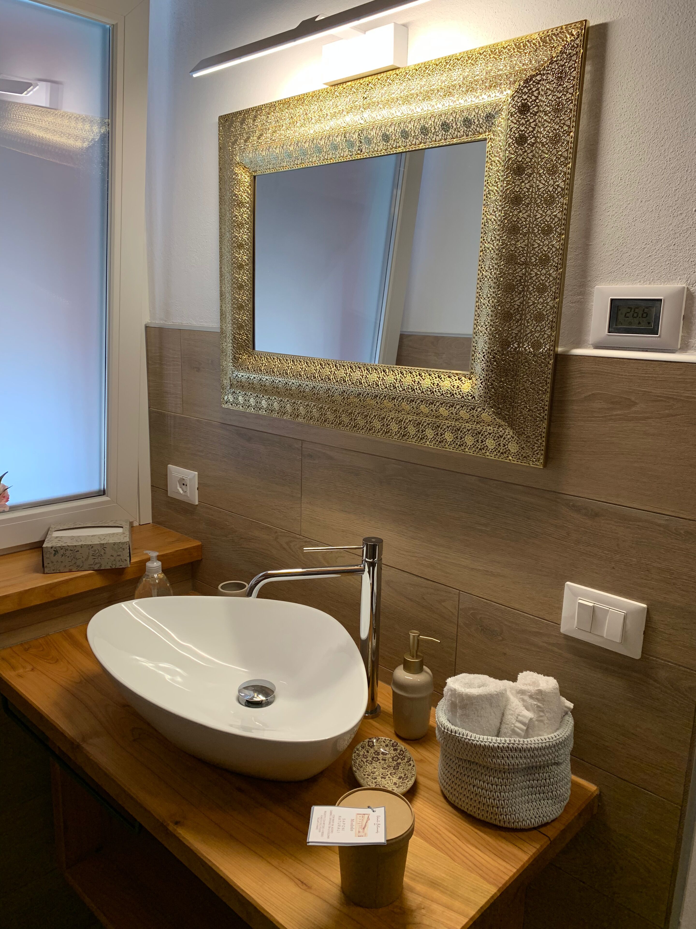 A bathroom with a white sink, a gold-framed mirror, a wooden vanity, a towel basket, and a digital thermostat.