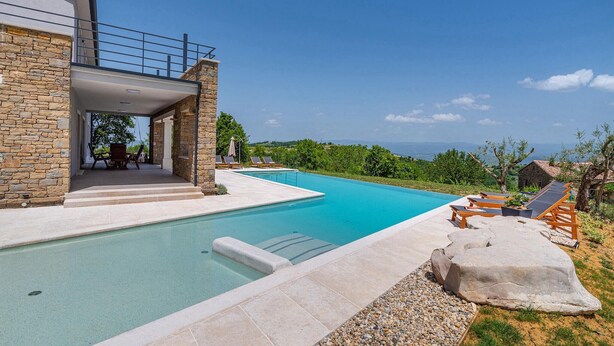Un espace moderne aménagé au bord de la piscine, avec une façade en pierre, un espace salon couvert et une vue dégagée sur le paysage.