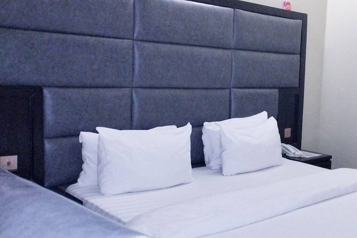 Hotellihuone, jossa on suuri sänky, kaksi valkoista tyynyä, yöpöytä, jossa on puhelin, ja seinälle asennettu televisio.