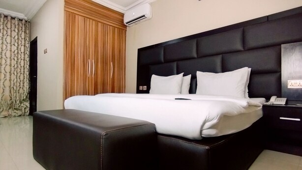 A neatly made bed with a black headboard, a bedside table with a phone, and a wooden panel wall. - Breakfort Hotel and Suites (Uyo)