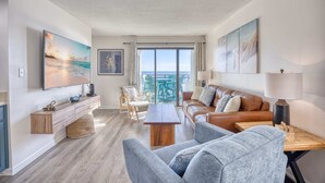 Un salon moderne avec un canapé, des fauteuils, une table basse et un téléviseur. Il y a un balcon avec vue sur la mer.