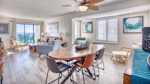 Un salon spacieux avec une table à manger en bois, des sièges confortables, un ventilateur de plafond et de grandes fenêtres équipées de stores.