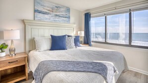 Une chambre avec un grand lit, une table de chevet avec une lampe, une fenêtre donnant sur l’océan et un tableau accroché au mur.