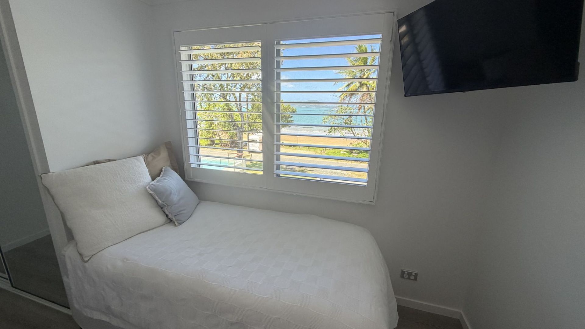 A single bed with a white mattress and pillows, a window with horizontal blinds, and a flat-screen TV mounted on the wall.