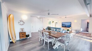 A modern living room with a dining area, a large flat-screen TV, and a surfboard decoration.