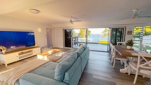 A modern living room with a large flat-screen TV, a sofa, a dining table with chairs, and a view of the outdoors. - Bella Luna Beach Front 2 bed (South Mission Beach)