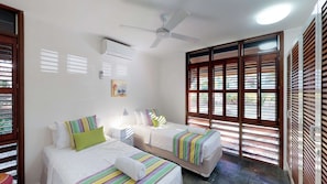 Quarto de hotel com duas camas, um ventilador de teto, aparelho de ar condicionado e janelas amplas com venezianas de madeira.