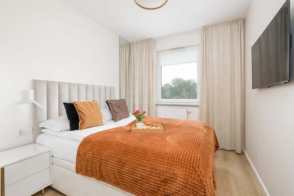 Un dormitorio moderno con cama, mesita de noche, televisor montado en la pared y ventana con cortinas.