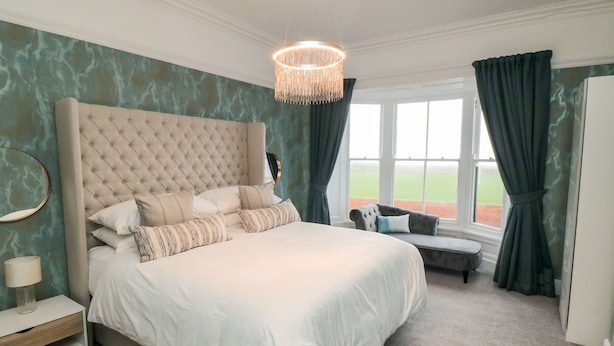 A bedroom with a large bed, a bedside table, a bench, and a window with curtains.