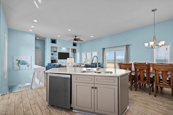 A modern kitchen with a central island, a dining area with wooden chairs, and a view of the ocean through large windows. - Risky Business #152, Oceanfront House in Topsail Beach, Pet Friendly! (Topsail Beach)