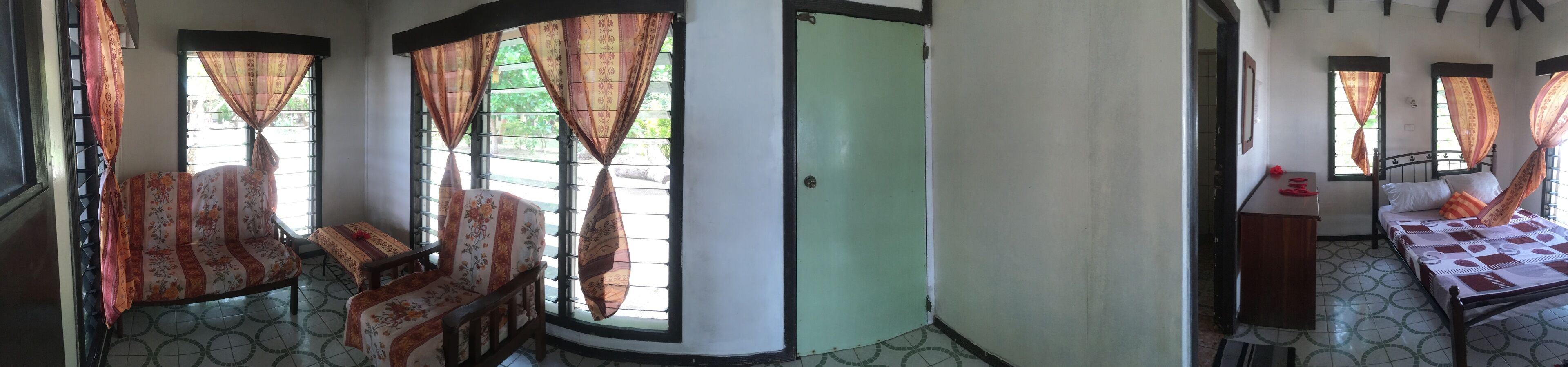 Ein Zimmer mit einem Bett, einem kleinen Tisch und einem Stuhl. Es gibt Fenster mit Vorhängen.