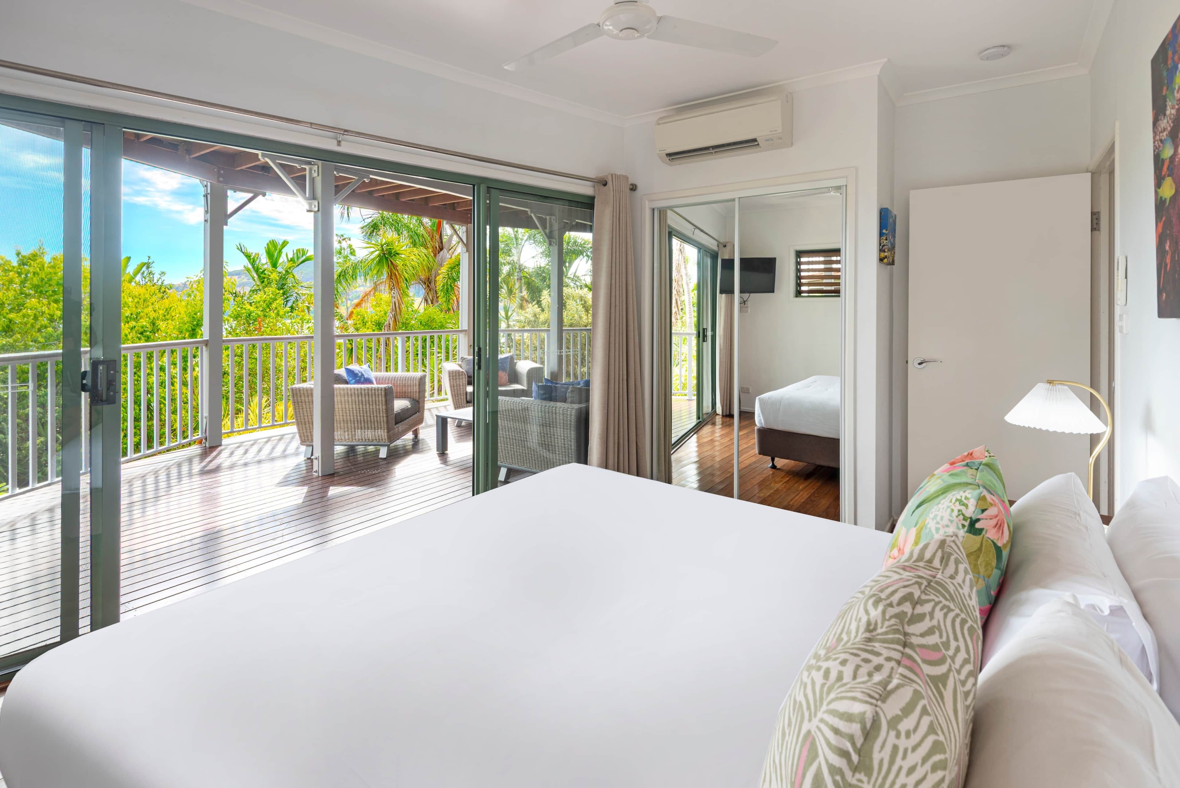 A bedroom with a large bed, a balcony with outdoor furniture, and a view of greenery.