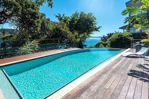 A swimming pool with a wooden deck, surrounded by trees and overlooking a body of water.