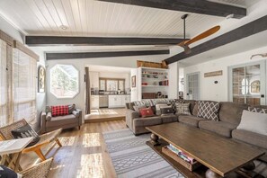 A spacious living area with a sofa, coffee table, and wicker chairs. There is a kitchen in the background with white cabinets and a dining area.