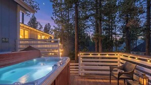 Un jacuzzi en una terraza de madera con luces colgantes, rodeado de árboles y un edificio con ventanas iluminadas.