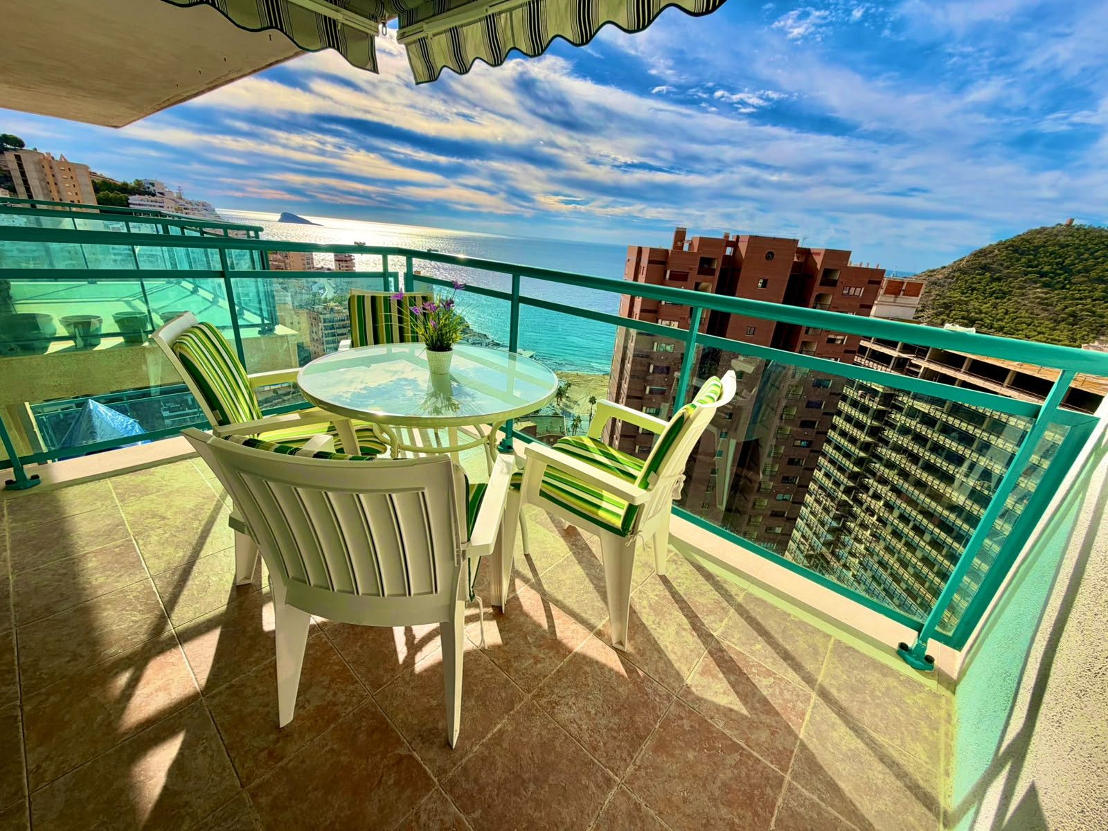 A balcony with a glass table and chairs, overlooking a coastal area with buildings and a hill.