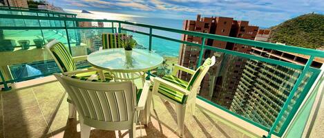 A balcony with a glass table and chairs, overlooking a coastal area with buildings and a hill.
