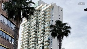 Un grand immeuble résidentiel doté de nombreux balcons, de palmiers et d’une voiture blanche garée devant.